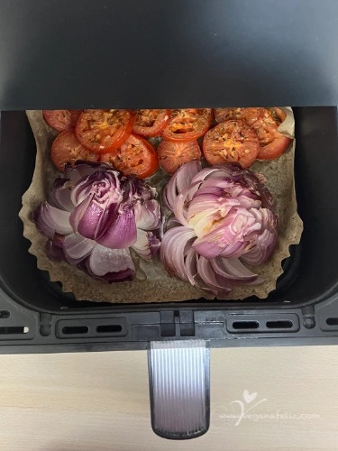 CebollaAirfryer Cebolla cortada en forma de flor y rodajas de tomate en la cesta de una airfryer antes de cocinar.
